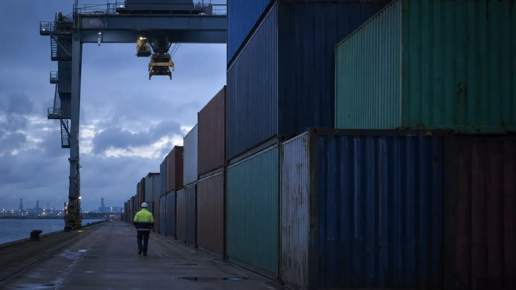 Stacked shipping containers on a European quayside at dusk with a gantry crane above and a lone inspector walking the row